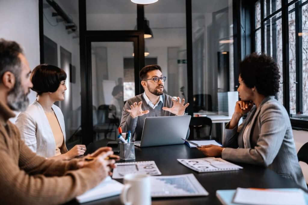 Vier Personen in Businesskleidung sitzen im Büro um einen Tisch und führen eine Nummer 1 Diskussion mit Laptops und Dokumenten